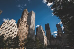A low angle shot captures a cluster of New York City skyscrapers against a vibrant blue sky dotted with fluffy white clouds, framed by tree foliage in the foreground.