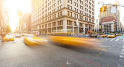 Busy road intersection in Manhattan, New York, at sunset with blurred yellow cabs on foreground, and a city building behind it.